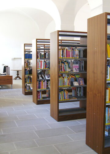 Ein Raum der Füssener Stadtbibliothek mit Regalen voller Bücher und einem Schreibtisch ist abgebildet. Die Wände sind hell und die Decke weist eine architektonische Struktur auf.