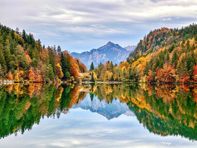 Im Fokus des Bildes liegt der Alatsee, dessen Oberfläche, glatt wie ein Spiegel, die umliegende Natur widerspiegelt. 