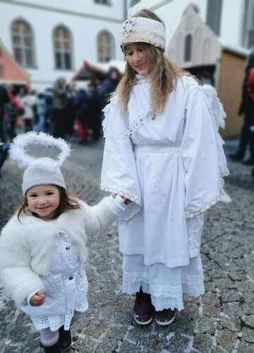 Das Bild fängt eine herzerwärmende Szene einer jungen Mädchen und einer Frau ein, beide gekleidet in Weiß, die zusammen auf einer Kopfsteinpflasterstraße spazieren gehen. Das Mädchen, welches auf der linken Seite ist, hält die Hand der Frau, was auf eine enge Beziehung zwischen ihnen hinweist. Die Frau, die auf der rechten Seite ist, trägt ein weißes Kleid und eine weiße Hut mit einem Federschmuck, sie verleiht ihrem Outfit damit eine elegante Note. Die Kopfsteinpflasterstraße, auf der sie gemeinsam gehen, ist von Gebäuden umgeben, was eine malerische Kulisse für diesen Moment schafft. Die Gesamtatmosphäre des Bildes ist von Wärme, Zusammengehörigkeit und Freude geprägt.