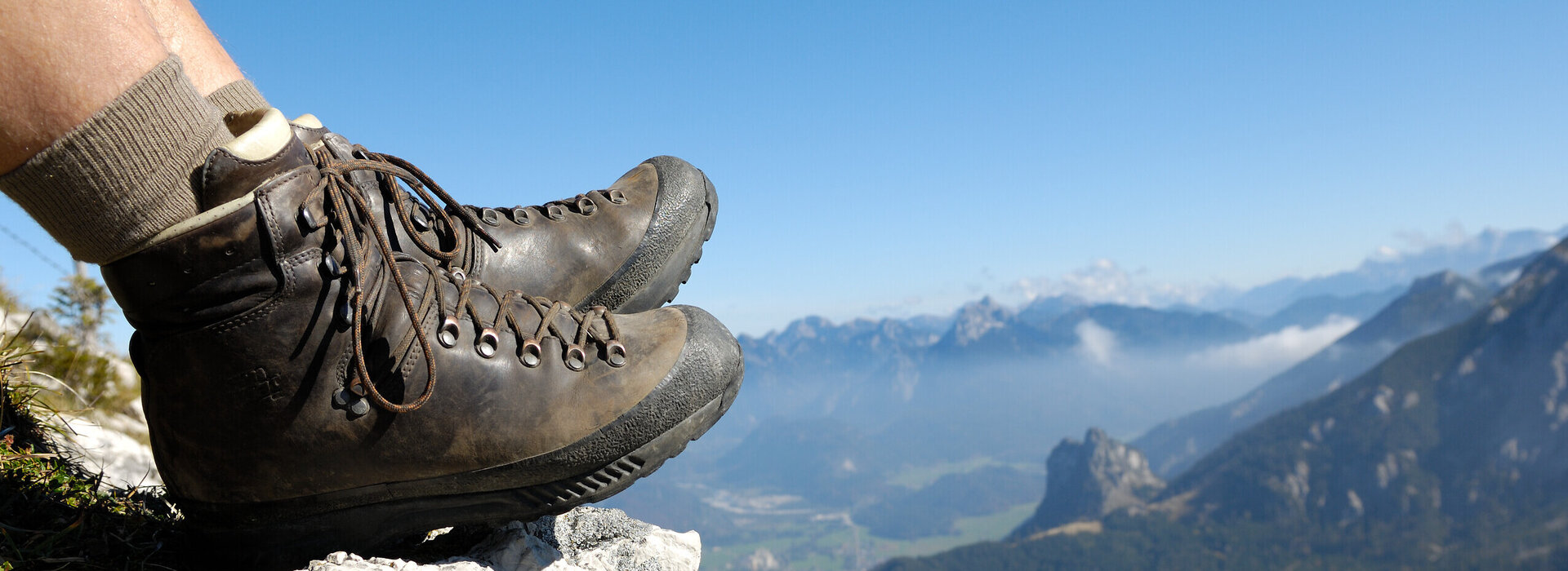 Das Bild zeigt die ledernen Bergschuhe einer Person, die auf einem Berg sitzt. Weiter unten erkennt man Bergwiesen, Wälder und ein Tal. Dahinter erheben sich weitere Berge. 