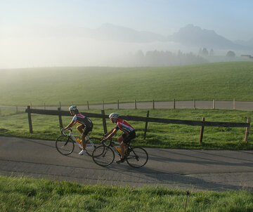 Das Bild zeigt zwei Rennradfahrer in Trikots und Radlerhosen, die einen Hügel emporfahren. Der Morgennebel lichtet sich langsam. Im Hintergrund erheben sich die Berge.  