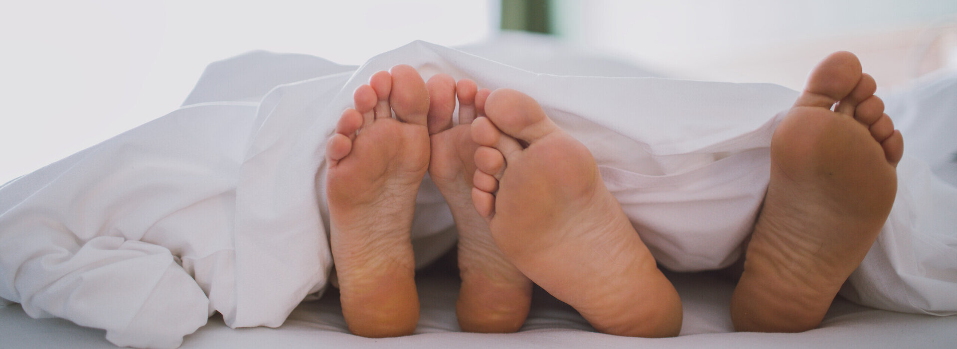 Das Bild zeigt zwei nackte Fußpaare, die unter einer weißen Decke hervorschauen. Das Bild suggeriert einen Moment der Entspannung und des Wohlbehagens.