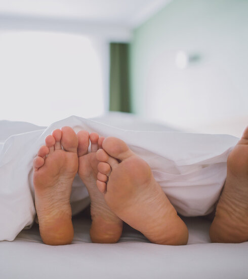 Das Bild zeigt zwei nackte Fußpaare, die unter einer weißen Decke hervorschauen. Das Bild suggeriert einen Moment der Entspannung und des Wohlbehagens.