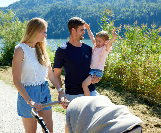 Eine junge Familie ist bei schönem Wetter mit dem Kinderwagen auf dem Rundweg am Hopfensee untwegs.