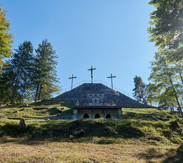 Das Bild zeigt drei Kreuze, die auf einer steinernen Aussichtsplattform stehen. Zu der Plattform führen zahlreiche Stufen hinauf. Ringsherum stehen große, grüne Bäume, durch deren Laub die Sonne scheint.