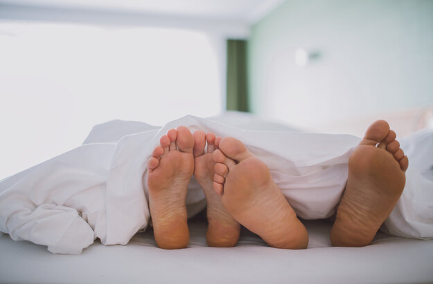 Das Bild zeigt zwei nackte Fußpaare, die unter einer weißen Decke hervorschauen. Das Bild suggeriert einen Moment der Entspannung und des Wohlbehagens.