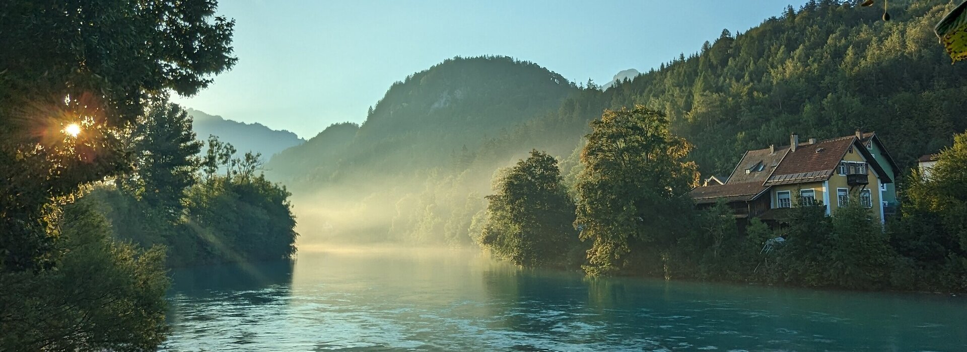 Das Bild zeigt einen Flusslauf mit blaugrünem Wasser, das von Bäumen und Hügeln gesäumt wird. Nebel liegt über dem Wasser und den Hügeln.