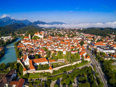 Das Foto zeigt Füssen von oben. Den Blick des Betrachters wird über die Dächer von Füssen geleitet. Im Vordergrund ist das Franziskanerkloster zu erkennen, das aus Klostergebäude und Klosterkirche St. Stephan besteht. Das Klostergelände ist eingerahmt von der historischen Stadtmauer. Neben dem Kloster fließt der türkisblau Fluss Lech. Im Hintergrund des Klosters erstreckt sich die Füssener Altstadt mit dem Hohen Schloss und dem Bergpanorama.