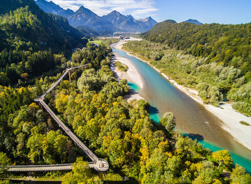 Das Foto zeigt das Flussbett des türkisblauen Flusses Lech, das von Kiesbänken gesäumt ist. Der Fluss ist von Wäldern flankiert, die in unterschiedlichen Grüntönen zu sehen sind. Durch den Wald am linken Flussufer zieht sich ein auf meterhohen Pfählen befestigter Holzweg – der Baumkronenweg, der im Zickzack verläuft. Das Foto wurden von oben aufgenommen. Im Hintergrund erheben sich die Berge. Der Himmel ist blau und von wenigen weißen Wolken durchzogen. 