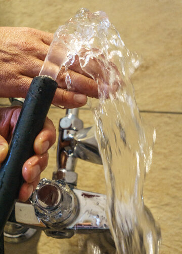 Das Bild zeigt zwei Hände die einen Wasserschlauch halten. Aus dem Wasserschlauch kommt ein klares Wasser, das über die eine Hand läuft. 