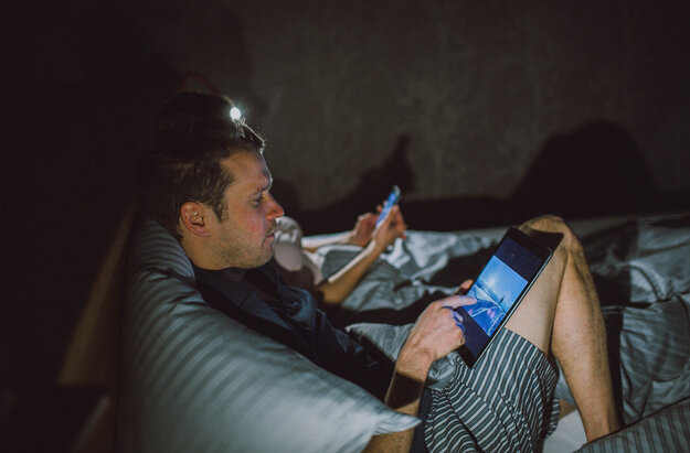 Das Bild zeigt zwei Personen, die nachts mit Handy und Tablet im Bett liegen. Sie lehnen nebeneinander am Kopfende. 