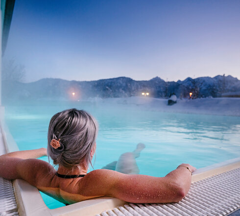 Das Bild zeigt eine Frau, die in Badekleidung in einem dampfenden Outdoor-Pool liegt. Die Landschaft im Hintergrund ist tief verschneit.  