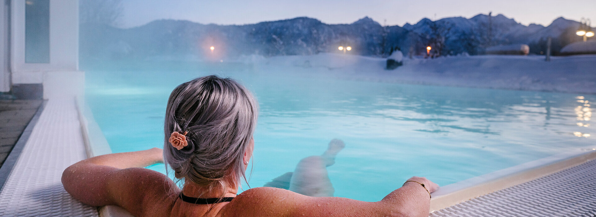 Das Bild zeigt eine Frau, die in Badekleidung in einem dampfenden Outdoor-Pool liegt. Die Landschaft im Hintergrund ist tief verschneit.  