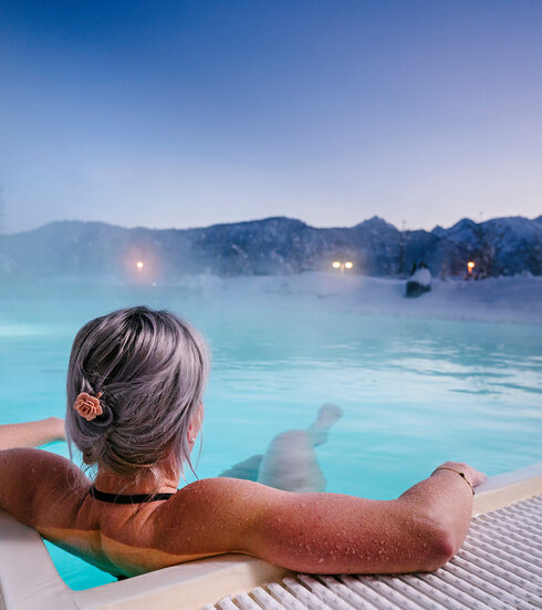 Das Bild zeigt eine Frau, die in Badekleidung in einem dampfenden Outdoor-Pool liegt. Die Landschaft im Hintergrund ist tief verschneit.  