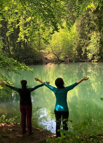 Das Bild zeigt zwei Frauen, die mit ausgebreiteten Armen mit dem Rücken zum Betrachter an einem kleinen See stehen. Den See und die Frauen umgibt ein dichter, grüner Laubwald.