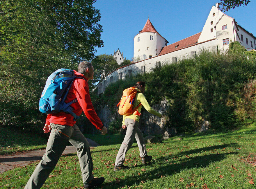 Das Bild zeigt einen Mann und eine Frau in Wanderkleidung und Rucksack, die eine Wiese entlang gehen. Über ihnen thront auf einem Felsen das Hohe Schloss. Im Hintergrund wachsen hohe grüne Bäume. Der Himmel ist blau. Die Sonne scheint.  