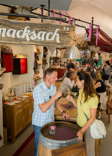 Ein Mann und eine Frau stehen vor einem Fass Wein und probieren Wein in einem rustikalen, gewölbten Raum. Der Raum ist mit Weinregalen, Ständen und anderen Produkten gefüllt.