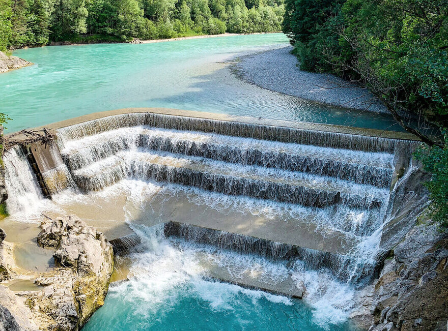 Das Bild zeigt einen künstilich angelegten Wasserfall, den Lechfall in Füssen. Im Hintergreund ist der Fluss Lech und Bäume im Sonnenschein unter blauem Himmel zu erkennen. 