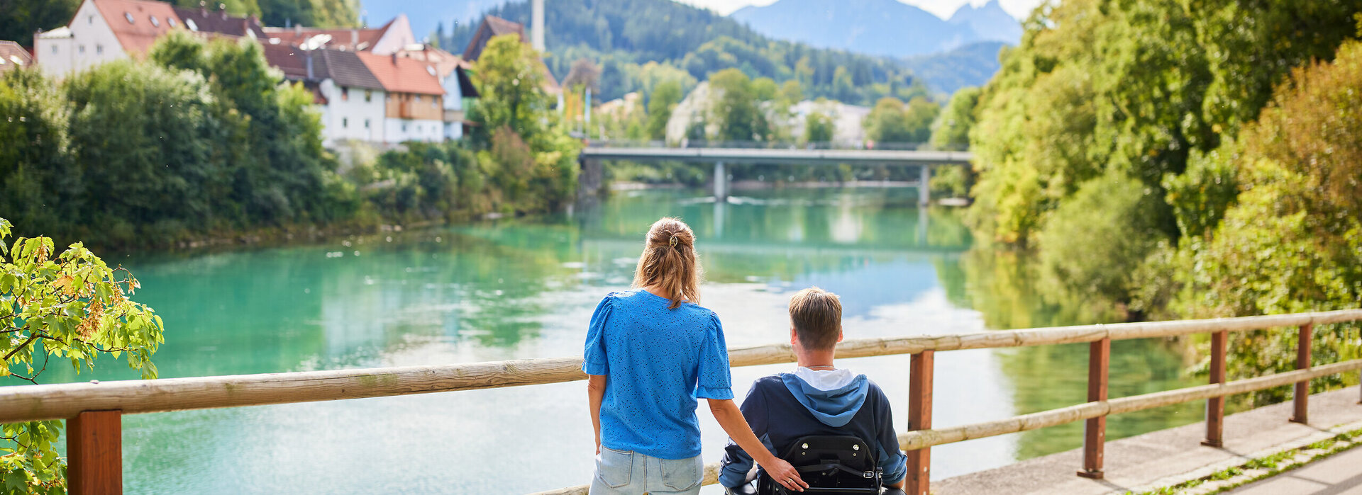 Das Bild fängt einen friedlichen Moment zweier Personen an einer Holzbrücke ein, die einen Blick auf eine Flusslandschaft bietet. Der Mann sitzt im Rollstuhl, während die Frau neben ihm steht. Im Hintergrund schmiegt sich ein malerisches Dorf in die üppige Vegetation. 