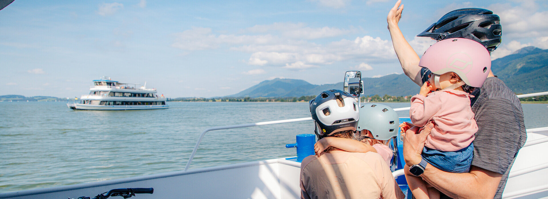 Das Bild zeigt eine friedliche Szene auf einem Boot. Eine Person mit einem pinken Helm hält ein Baby in den Armen. Das Baby trägt ebenfalls einen pinken Helm, was auf ein gemeinsames Interesse an Sicherheit hindeutet. Der Mann trägt außerdem einen pinken Helm, was darauf hindeuten könnte, dass er der Vater des Babys ist.  Das Boot, auf dem sie sich befinden, ist weiß und blau und liegt in einem Hafen. Der Hafen ist von Bergen umgeben, was der Szene einen Hauch von Natur verleiht. In der Ferne ist ein großes Boot zu sehen, was darauf hindeuten könnte, dass sie sich in einem beliebten Wassersportgebiet befinden.  Der Mann und das Baby sind nicht allein auf dem Boot. Auch ein Fahrrad ist vorhanden, was darauf hindeuten könnte, dass sie eine gemütliche Fahrt oder Exkursion unternehmen. Das Fahrrad steht auf dem Boot bereit, um verwendet zu werden, sobald sie ihr Ziel erreicht haben.  Insgesamt zeichnet das Bild das Bild einer Familie, die einen Tag auf dem Wasser genießt, mit Sicherheit an erster Stelle.