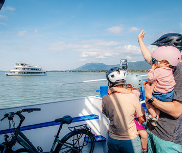 Das Bild zeigt eine friedliche Szene auf einem Boot. Eine Person mit einem pinken Helm hält ein Baby in den Armen. Das Baby trägt ebenfalls einen pinken Helm, was auf ein gemeinsames Interesse an Sicherheit hindeutet. Der Mann trägt außerdem einen pinken Helm, was darauf hindeuten könnte, dass er der Vater des Babys ist.  Das Boot, auf dem sie sich befinden, ist weiß und blau und liegt in einem Hafen. Der Hafen ist von Bergen umgeben, was der Szene einen Hauch von Natur verleiht. In der Ferne ist ein großes Boot zu sehen, was darauf hindeuten könnte, dass sie sich in einem beliebten Wassersportgebiet befinden.  Der Mann und das Baby sind nicht allein auf dem Boot. Auch ein Fahrrad ist vorhanden, was darauf hindeuten könnte, dass sie eine gemütliche Fahrt oder Exkursion unternehmen. Das Fahrrad steht auf dem Boot bereit, um verwendet zu werden, sobald sie ihr Ziel erreicht haben.  Insgesamt zeichnet das Bild das Bild einer Familie, die einen Tag auf dem Wasser genießt, mit Sicherheit an erster Stelle.