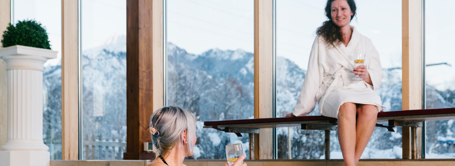 Das Bild zeigt einen Indoor-Swimming Pool. Zwei Frauen lächeln sich zu. Hinter einer großen Fensterfront erheben sich die schneebedeckten Berge.     
