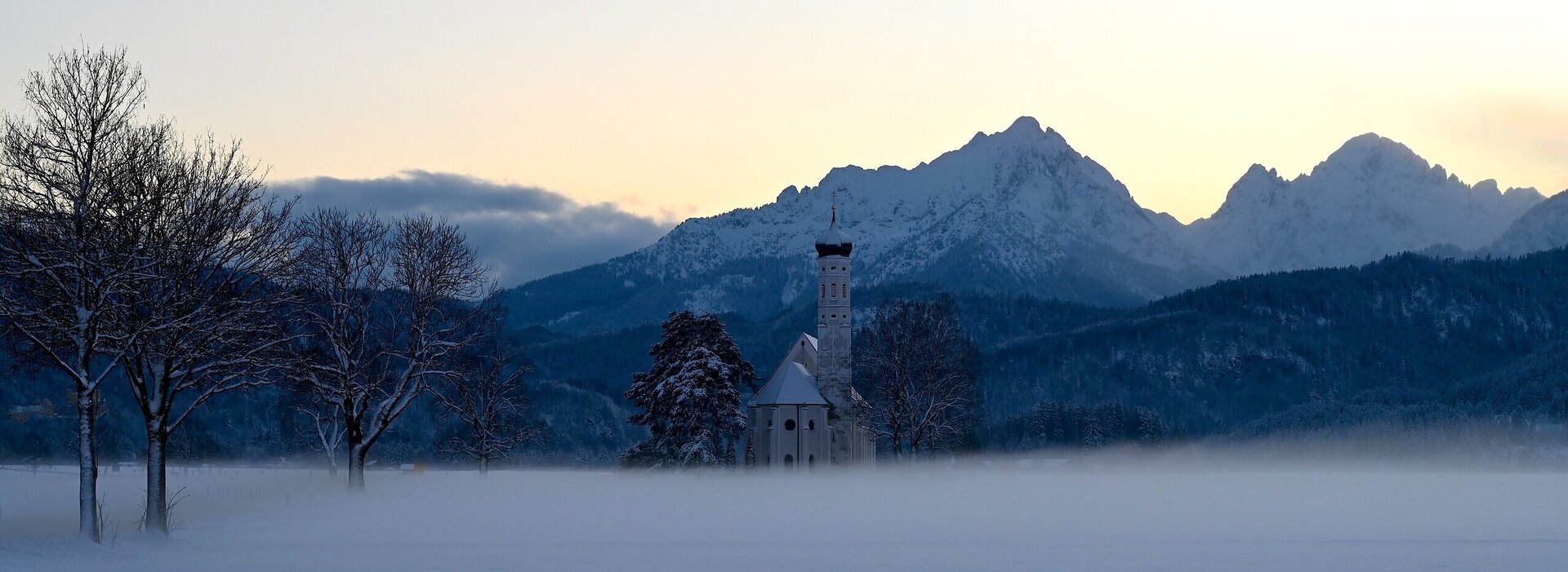 Das Bild zeigt eine Kirche in schneebedeckter und nebeliger Landschaft. Im Hintergrund bildet sich der Sonnenuntergang in der Bergkette. 