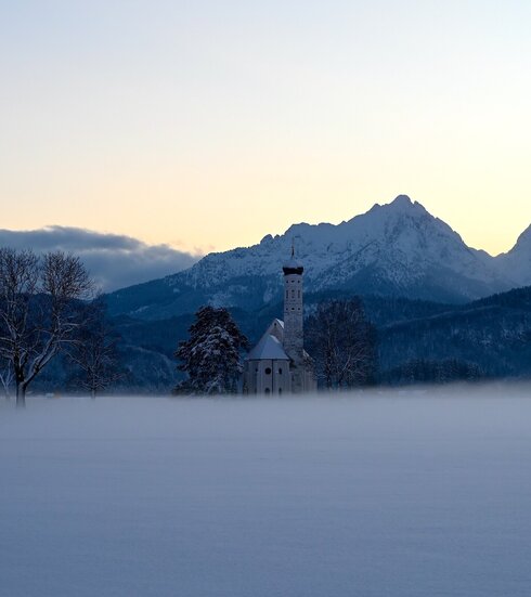 Das Bild zeigt eine Kirche in schneebedeckter und nebeliger Landschaft. Im Hintergrund bildet sich der Sonnenuntergang in der Bergkette. 