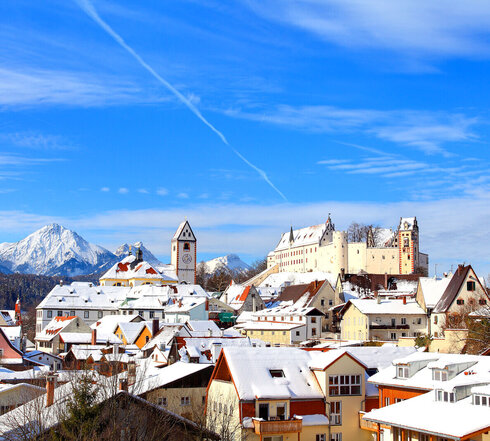 Das Bild zeigt die verschneite Füssener Altstadt mit dem imposanten Hohen Schloss im Hintergrund. Ein blauer Himmel mit Wolken und Flugstreifen bildet die Kulisse.
