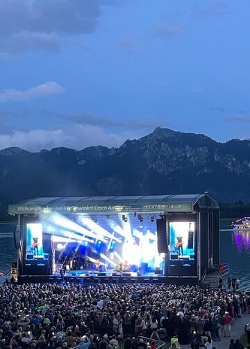 Das Foto zeigt ein Publikum vor einer Outdoor-Bühne mit Blick auf den Forggensee.