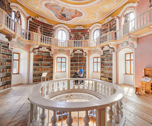 Die Bibliothek des Hohen Schlosses in Füssen mit hohen Regalen und einem beeindruckenden Rundbogen. Das Bild zeigt einen Raum voller Wissen und Geschichte.
