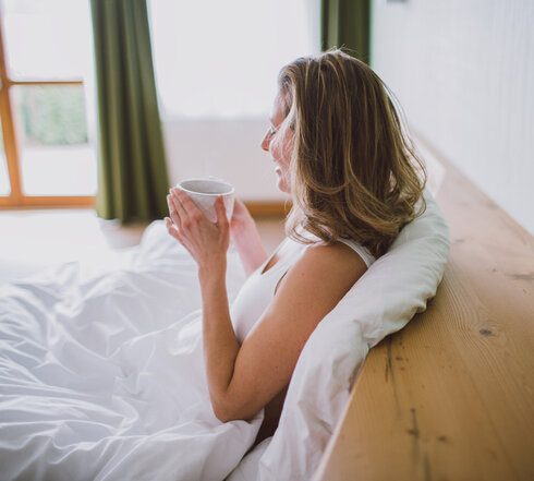 Das Bild zeigt eine Frau, die in einem Bett, das weiß bezogen ist, sitzt. Sie lehnt an der Rückwand des Bettes und hält eine Teetasse in der Hand.