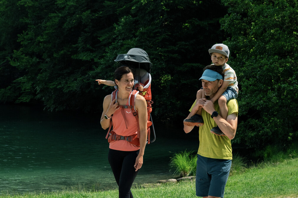 Eine Familie wandert auf einem Weg entlang eines Gewässers. Die Mutter trägt ein Kind im Tragegurt, während der Vater einen anderen Jungen auf den Schultern hat. Die Umgebung ist grün und natürliche, mit Bäumen im Hintergrund. Es ist ein sonniger Tag.