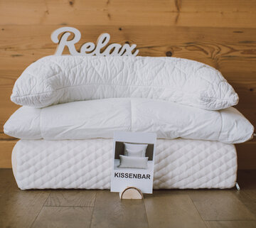 Das Bild zeigt drei unterschiedliche, aufeinanderliegende Kopfkissen, ohne Bezug, vor einer Holzwand. Auf dem obersten Kissen steht „Relax“ in Holzbuchstaben.
