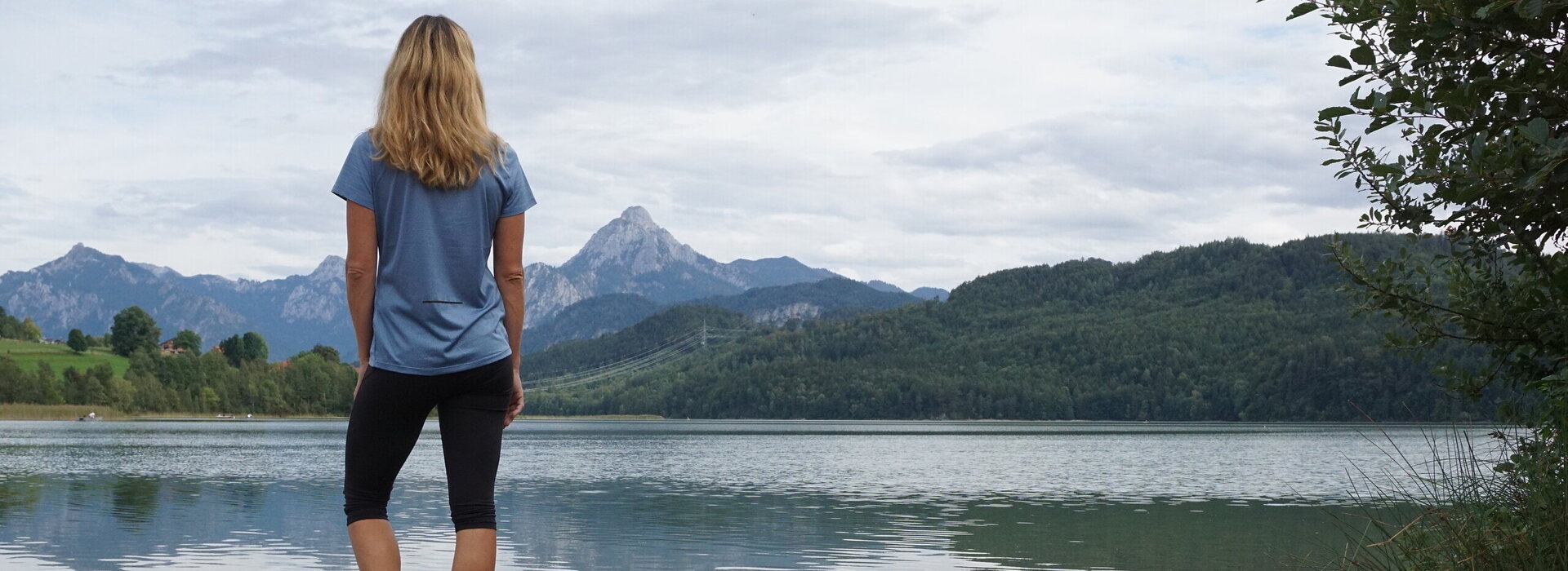 Ein See mit einem bewaldeten Ufer wird von einem Felsvorsprung und einer Person, die barfuß steht, dominiert. Im Hintergrund erheben sich Berge.