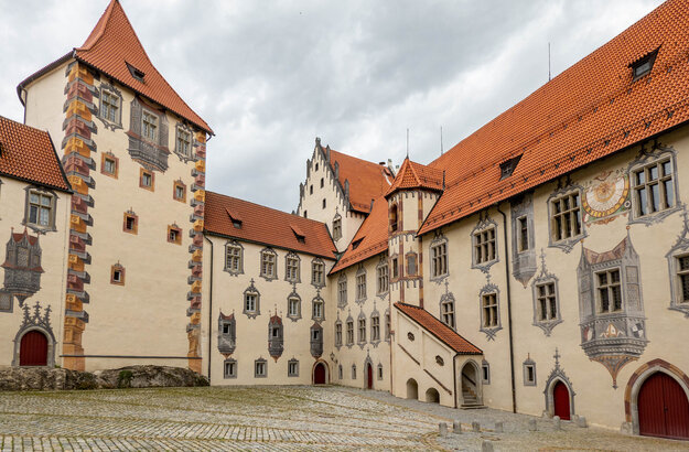 Das Bild zeigt den Innenhof des Hohen Schlosses in Füssen, der durch seine Illusionsmalerei an den Fassaden beeindruckt. Das Schlossgebäude hat rote Türen und ein Ziegeldach.