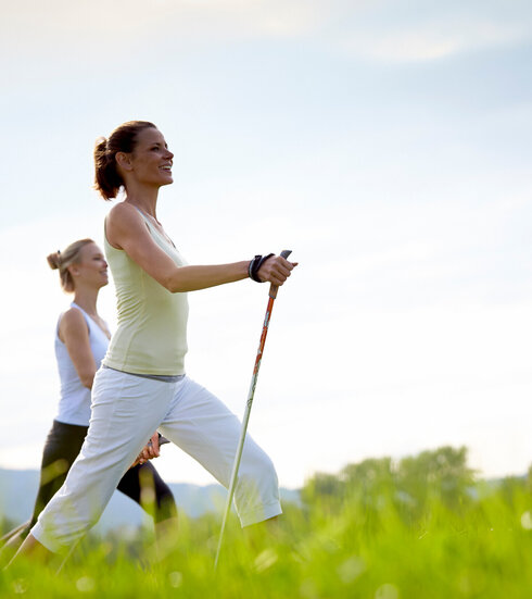 Das Bild zeigt zwei sportlich gekleidete, schlanke Frauen, die mit Nordic-Walking-Stöcken über eine grüne Wiese laufen. Der blaue Himmel ist von sanften Wolken durchzogen.