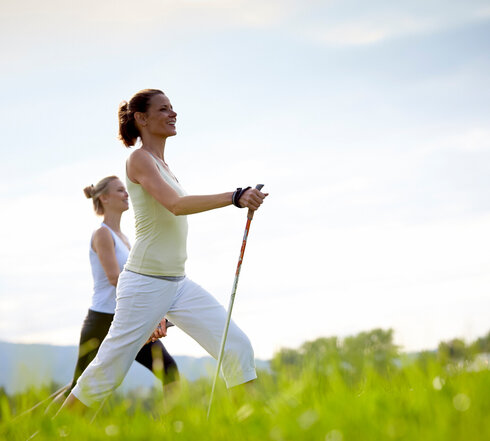 Das Bild zeigt zwei sportlich gekleidete, schlanke Frauen, die mit Nordic-Walking-Stöcken über eine grüne Wiese laufen. Der blaue Himmel ist von sanften Wolken durchzogen.
