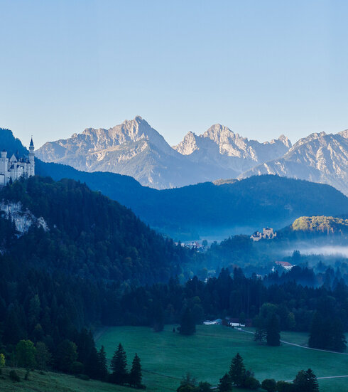 Das Bild zeigt die beiden Schlösser Hohenschwangau und Neuschwanstein, die auf zwei Anhöhen über den umliegenden Wiesen und Wäldern thronen. Im Hintergrund erheben sich die Berge. Der Himmel ist blau. Die Sonne geht gerade auf. Die Schlösser liegen noch im Schatten. 