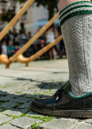 Das Bild zeigt mehrere Alphornspieler. Allerdings sind nur zwei Beine bis zu den Knien zu sehen. Die Beine sind mit grauen Kniestrümpfen und schwarzen Haferlschuhen bekleidet. Um die Alphörner kann man mehrere Zuschauer erkennen. Die Sonne scheint. Die Szenerie findet wohl auf einem Platz in der Stadt statt. Im Hintergrund sind Hausfassaden zu erkennen. Auf dem Boden liegt Kopfsteinpflaster.   