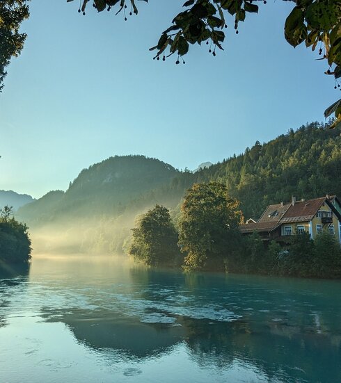 Das Bild zeigt einen Flusslauf mit blaugrünem Wasser, das von Bäumen und Hügeln gesäumt wird. Nebel liegt über dem Wasser und den Hügeln.
