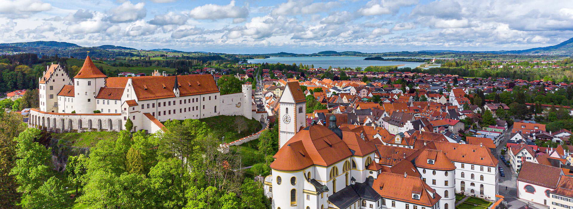 The picture offers a breathtaking view of the High Castle, the landmark of Füssen, Germany. The castle, a magnificent structure with a red roof, sits sublimely on a hill overlooking the town below. The town itself is a vibrant display of red roofs, creating an impressive contrast against the lush vegetation that surrounds it. The sky above is a clear blue, interspersed with fluffy white clouds that add to the overall serene and picturesque atmosphere of the scene. The perspective of the image is from a high view, providing a comprehensive view of the castle and the town below. The image conveys the essence of the castle and its surroundings, providing an insight into the rich history and architectural beauty of this famous landmark.