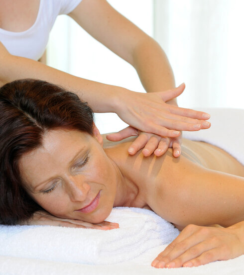 Das Bild zeigt eine Frau, die bäuchlings auf einer Massageliege liegt. Über ihr steht eine Person, die ihr die obere Rückenpartie massiert. 