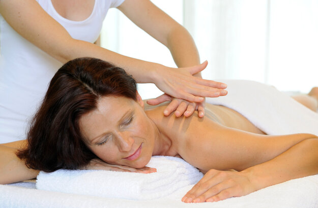 Das Bild zeigt eine Frau, die bäuchlings auf einer Massageliege liegt. Über ihr steht eine Person, die ihr die obere Rückenpartie massiert. 