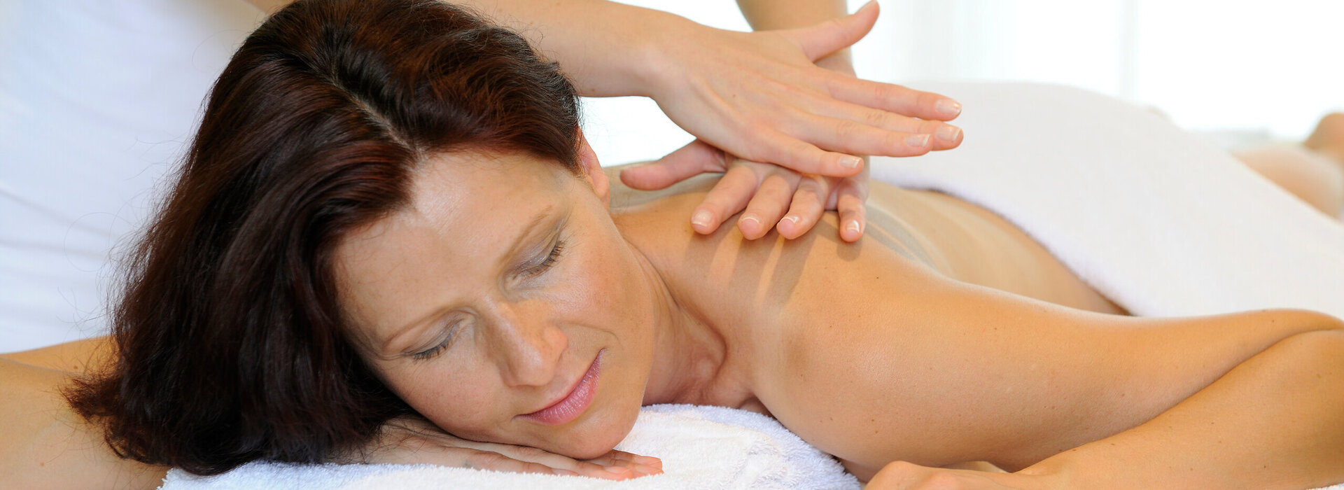 Das Bild zeigt eine Frau, die bäuchlings auf einer Massageliege liegt. Über ihr steht eine Person, die ihr die obere Rückenpartie massiert. 