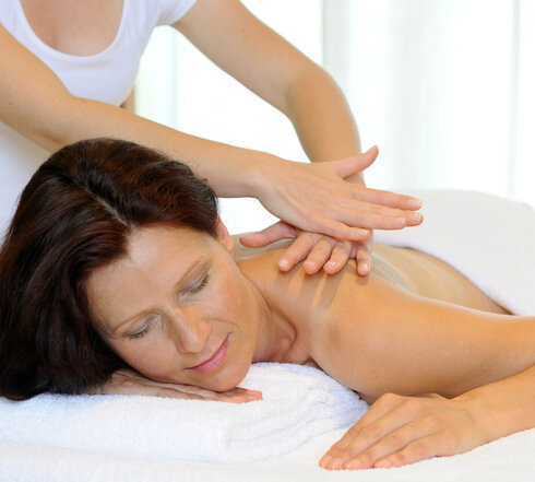 Das Bild zeigt eine Frau, die bäuchlings auf einer Massageliege liegt. Über ihr steht eine Person, die ihr die obere Rückenpartie massiert. 