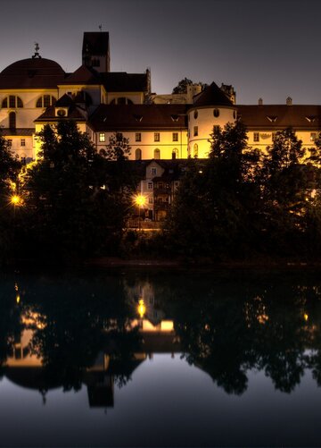 Das Hoch Schloss und der Klosterhof in Füssen erheben sich über den Lech, beleuchtet bei Nacht. Die Gebäude spiegeln sich im Fluss wider.