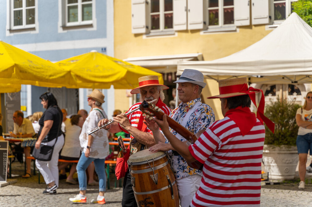 Musiker spielen auf einer belebten Gasse in der Füssener Altstadt. Die Darsteller tragen farbenfrohe Kleidung und sorgen mit Flöte und Trommel für Unterhaltung.            