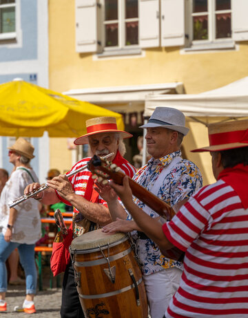 Musiker spielen auf einer belebten Gasse in der Füssener Altstadt. Die Darsteller tragen farbenfrohe Kleidung und sorgen mit Flöte und Trommel für Unterhaltung.            