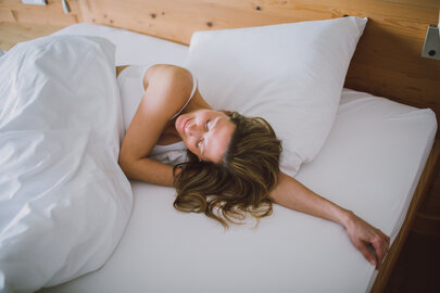 Das Bild zeigt eine Frau, die in einem Bett liegt, das weiß bezogen ist. Ihr Kopf liegt auf ihrem linken Arm. Sie hat die Augen geschlossen und wirkt entspannt.